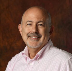 Photo of a smiling, elderly and bald white man with a brown moustache and grey beard wearing a white button-up shirt.