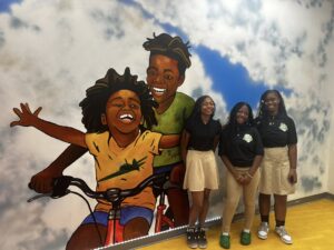 Three Black female students wearing school uniforms stand in front of a mural depicting children of color enjoying a bike ride.