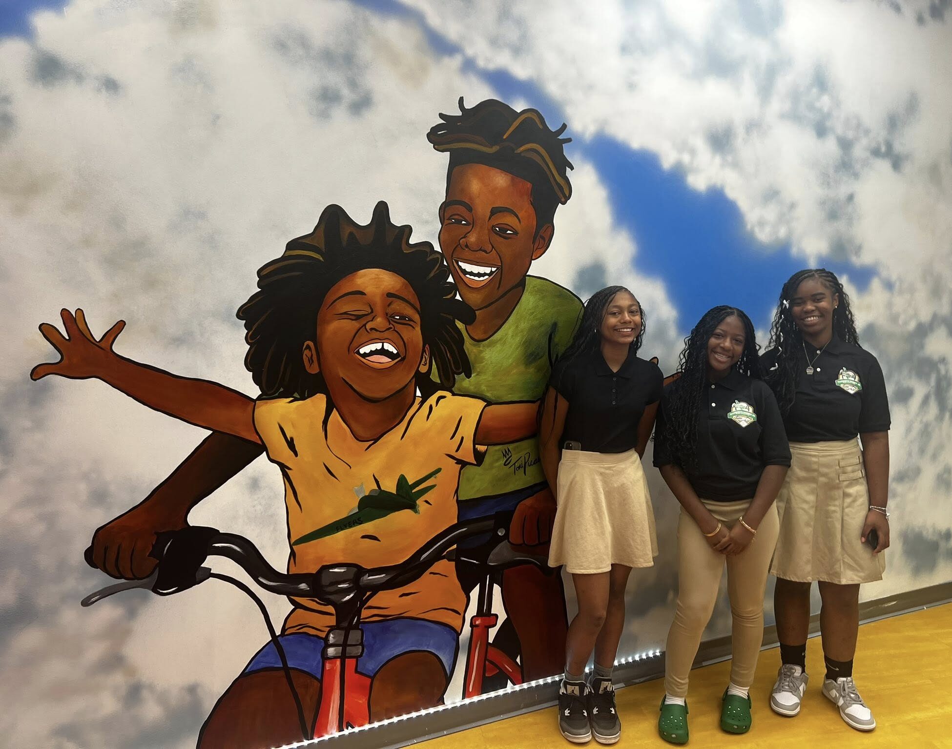 Three Black female students wearing school uniforms stand in front of a mural depicting children of color enjoying a bike ride.