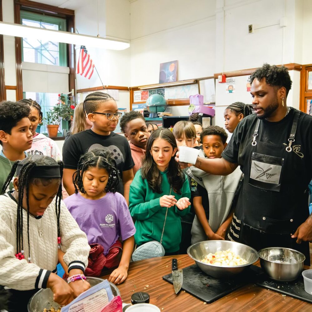 A group of school children and a Black man wearing an apron stand grouped around a table full of cooking implements.
